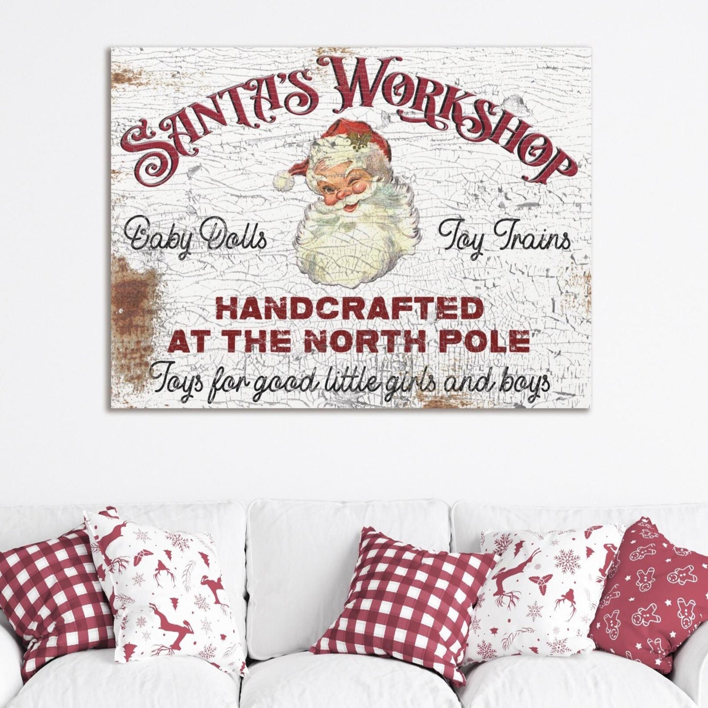 May include: A vintage white canvas sign with red text that reads 'Santa's Workshop' with a picture of Santa Claus. The sign also reads 'Baby Dolls', 'Toy Trains', 'Handcrafted at the North Pole', and 'Toys for good little girls and boys'. The sign is hanging on a white wall above a white couch with red and white checkered pillows.