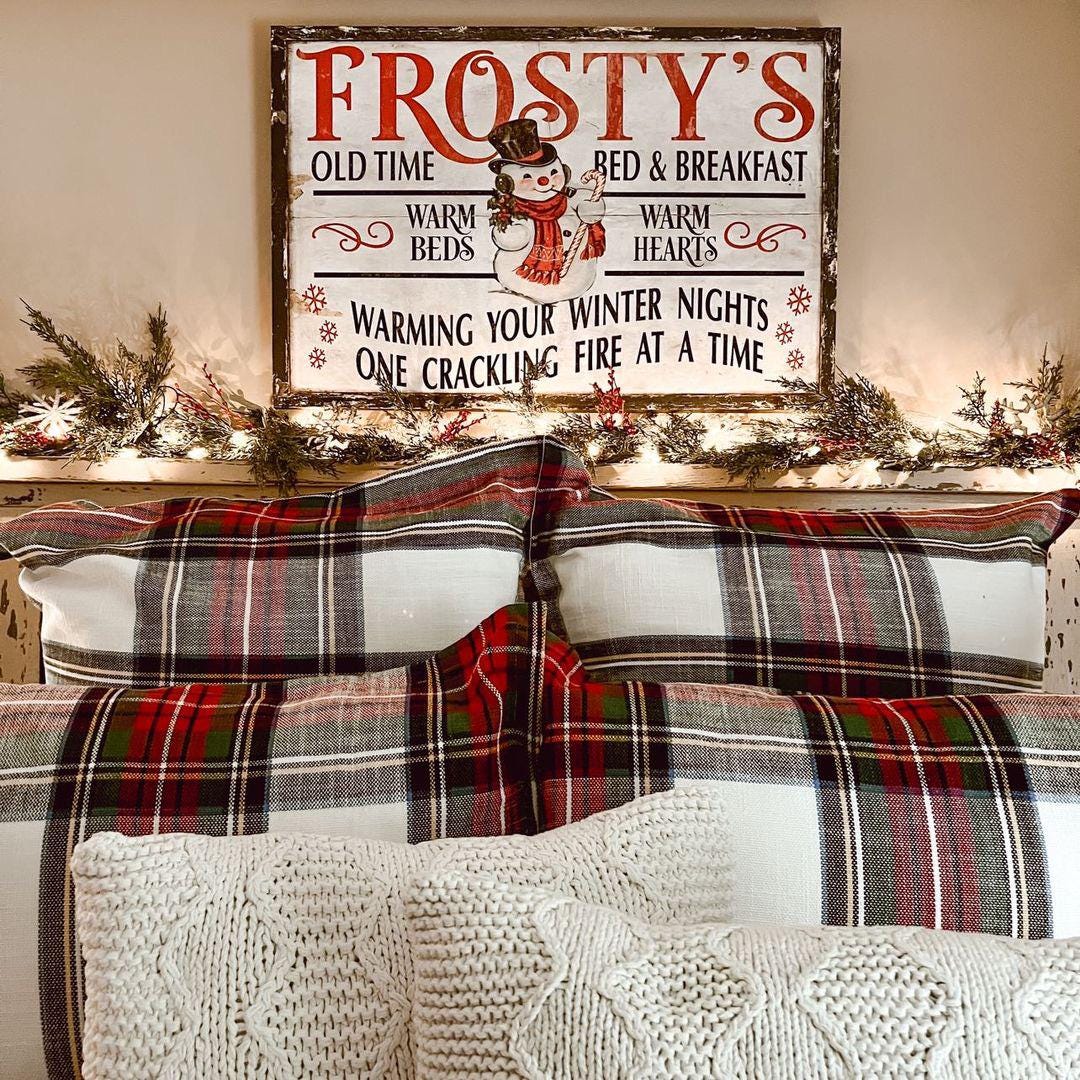 May include: Red and green plaid bedding on a bed with cream-colored knit throw pillows. Above the bed is a rustic wood sign that reads 'Frosty's Old Time Bed & Breakfast,' with a snowman graphic and the text 'Warming your winter nights, one crackling fire at a time.' The sign has a vintage design with red and white lettering.