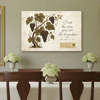 A dining room scene with a dark wood dining table and chairs covered in a white tablecloth, set on a patterned area rug against sage green walls. A framed botanical wall art print hangs above the table, flanked by two gold-framed mirrors.
