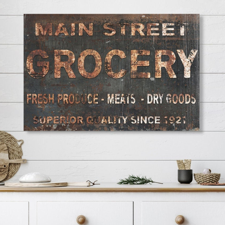 May include: A rustic, black and brown sign with white lettering that reads &#39;Main Street Grocery&#39;, &#39;Fresh Produce - Meats - Dry Goods&#39;, and &#39;Superior Quality Since 1921&#39;. The sign is hanging on a white wood wall above a white cabinet with two drawers.
