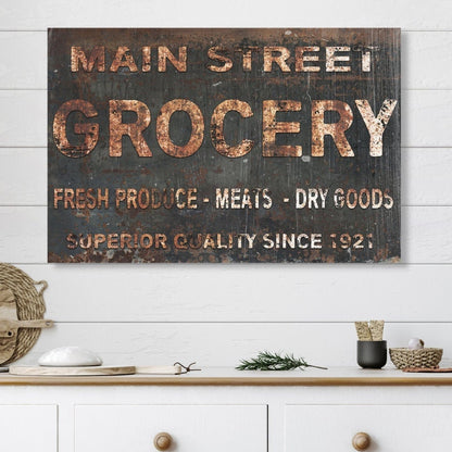 May include: A rustic, black and brown sign with white lettering that reads &#39;Main Street Grocery&#39;, &#39;Fresh Produce - Meats - Dry Goods&#39;, and &#39;Superior Quality Since 1921&#39;. The sign is hanging on a white wood wall above a white cabinet with two drawers.