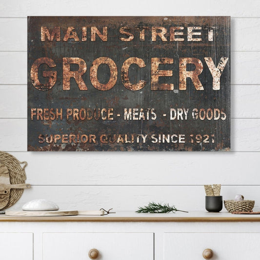 May include: A rustic, black and brown sign with white lettering that reads &#39;Main Street Grocery&#39;, &#39;Fresh Produce - Meats - Dry Goods&#39;, and &#39;Superior Quality Since 1921&#39;. The sign is hanging on a white wood wall above a white cabinet with two drawers.