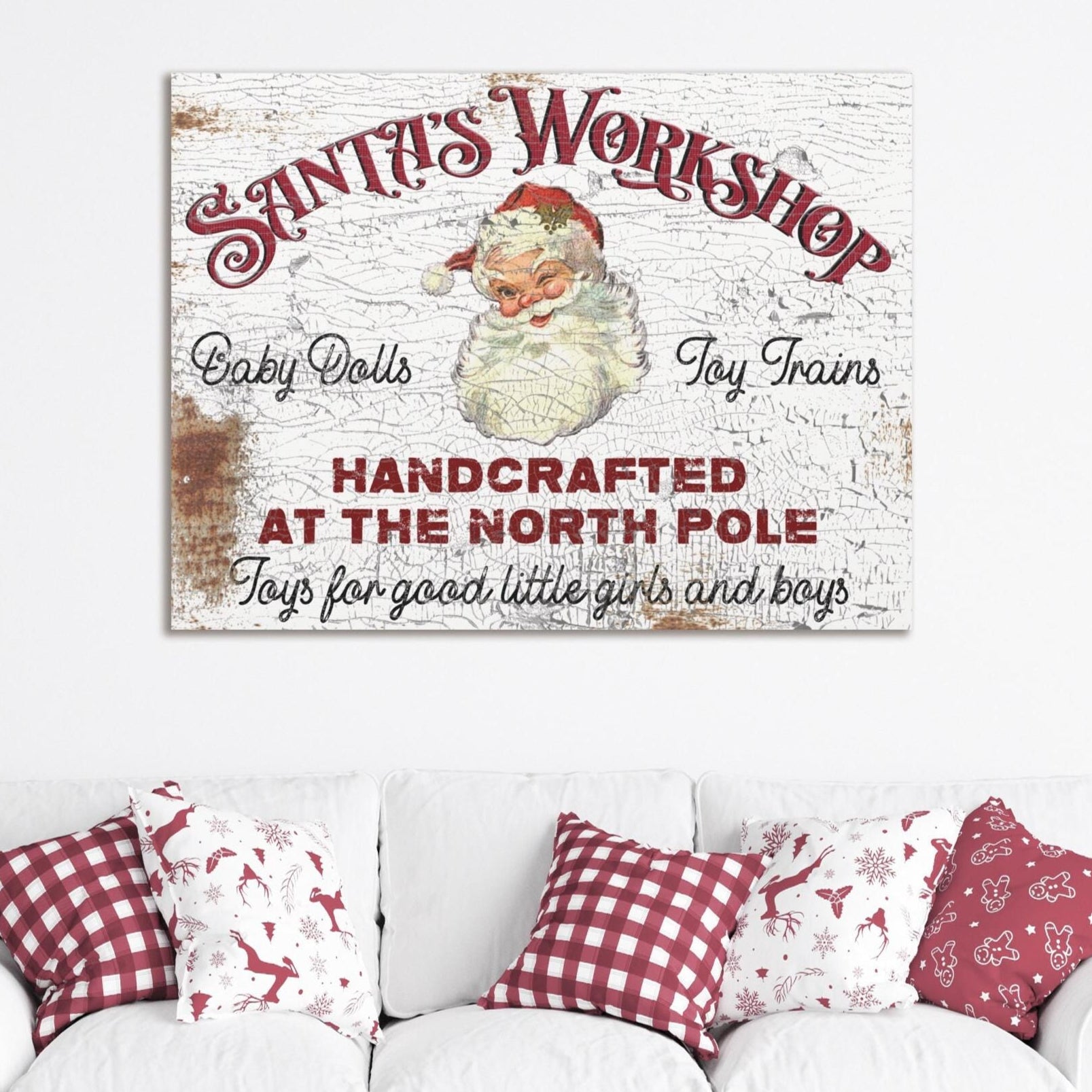 May include: A vintage white canvas sign with red text that reads &#39;Santa&#39;s Workshop&#39; with a picture of Santa Claus. The sign also reads &#39;Baby Dolls&#39;, &#39;Toy Trains&#39;, &#39;Handcrafted at the North Pole&#39;, and &#39;Toys for good little girls and boys&#39;. The sign is hanging on a white wall above a white couch with red and white checkered pillows.