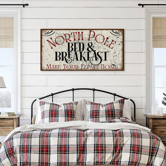 May include: A black metal bed frame with a white and red plaid comforter and matching pillows. A sign above the bed reads &#39;North Pole Bed & Breakfast Make Your Elf At Home&#39; in black and red lettering on a white background.