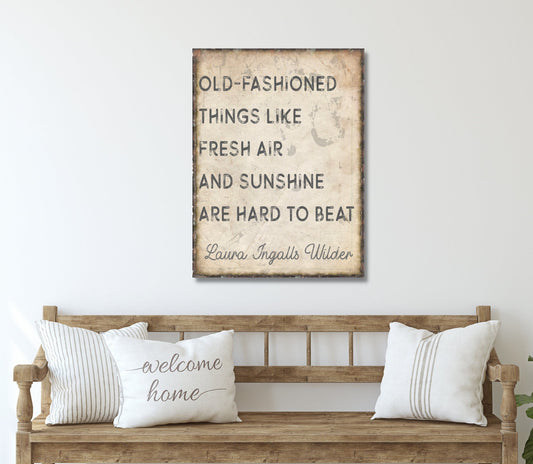 May include: A canvas print with a quote by Laura Ingalls Wilder that reads &#39;Old-fashioned things like fresh air and sunshine are hard to beat.&#39; The print is on a beige background with a distressed look and is hanging on a white wall above a wooden bench with two pillows. One pillow has the words &#39;welcome home&#39; written on it in cursive.