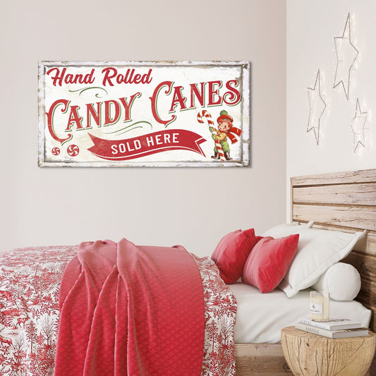 May include: A vintage-style sign with red and white lettering that reads &#39;Hand Rolled Candy Canes Sold Here&#39;. The sign has a distressed white background and features a cartoon image of a person holding a candy cane. The sign is hanging on a white wall above a bed with red and white bedding.