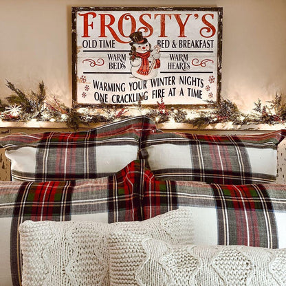 May include: Red and green plaid bedding on a bed with cream-colored knit throw pillows. Above the bed is a rustic wood sign that reads &#39;Frosty&#39;s Old Time Bed & Breakfast,&#39; with a snowman graphic and the text &#39;Warming your winter nights, one crackling fire at a time.&#39; The sign has a vintage design with red and white lettering.