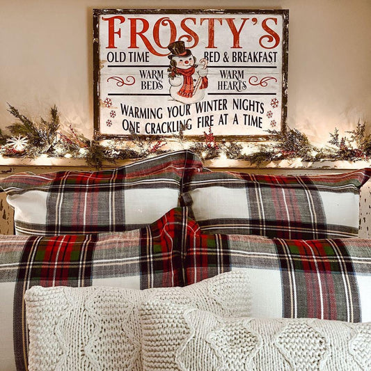 May include: Red and green plaid bedding on a bed with cream-colored knit throw pillows. Above the bed is a rustic wood sign that reads &#39;Frosty&#39;s Old Time Bed & Breakfast,&#39; with a snowman graphic and the text &#39;Warming your winter nights, one crackling fire at a time.&#39; The sign has a vintage design with red and white lettering.